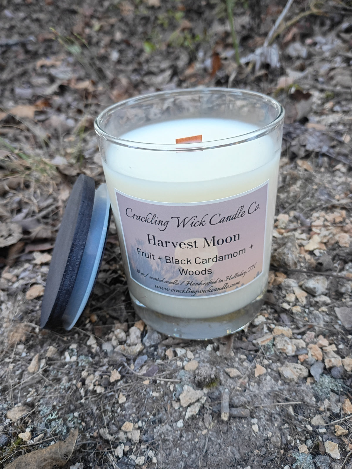 Scented candle in clear glass jar with cream wax and wooden wick; lid beside